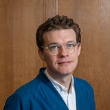 Photograph of a white male in his early forties with short sandy hair. He is smiling and wearing a blue cardigan over a white shirt.